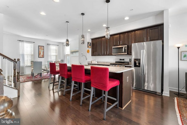 kitchen with appliances with stainless steel finishes, hanging light fixtures, a kitchen island with sink, dark brown cabinetry, and dark wood-type flooring