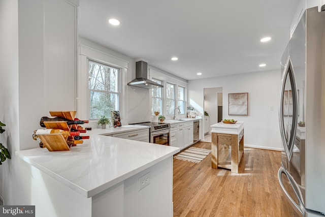 kitchen with appliances with stainless steel finishes, white cabinets, kitchen peninsula, wall chimney range hood, and light wood-type flooring