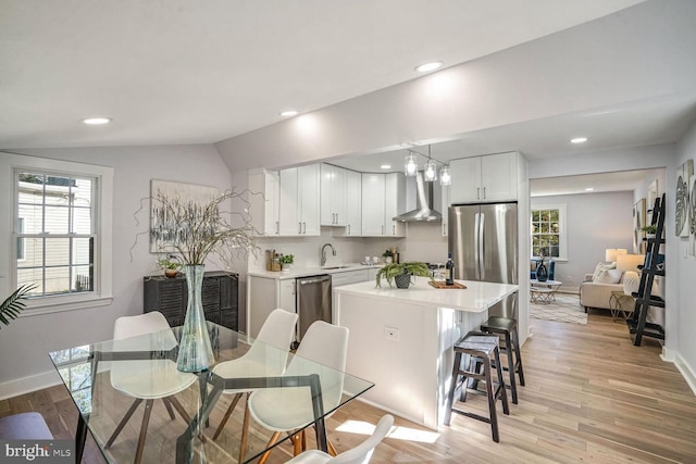 kitchen featuring wall chimney range hood, white cabinetry, hanging light fixtures, stainless steel appliances, and light hardwood / wood-style floors