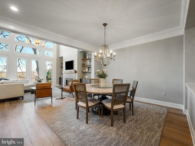 dining room featuring hardwood / wood-style floors, baseboards, a warm lit fireplace, ornamental molding, and a chandelier