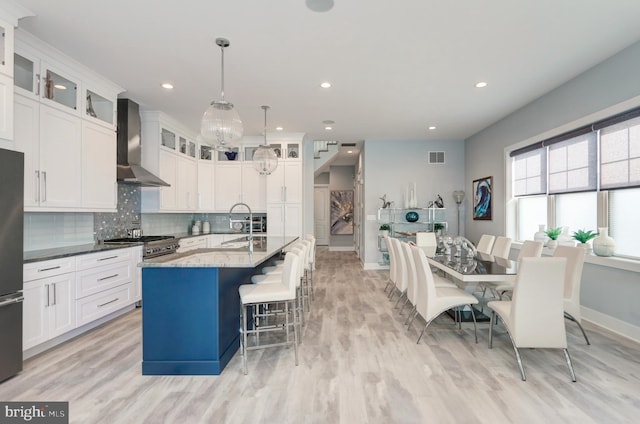 kitchen featuring wall chimney range hood, decorative backsplash, a center island with sink, decorative light fixtures, and dark stone counters