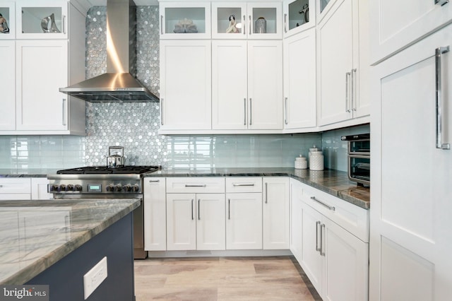 kitchen featuring wall chimney range hood, high end stove, white cabinets, decorative backsplash, and dark stone counters