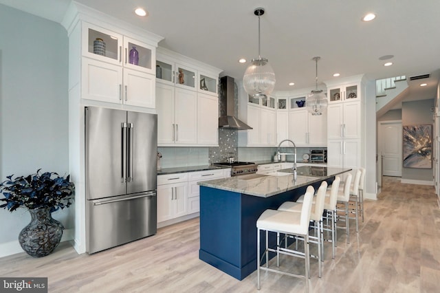 kitchen with wall chimney exhaust hood, white cabinetry, high end refrigerator, an island with sink, and dark stone counters