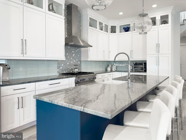 kitchen featuring a kitchen island with sink, wall chimney range hood, stainless steel range, and dark stone counters