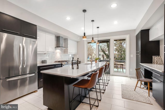 kitchen with wall chimney range hood, stainless steel fridge, a breakfast bar, white cabinetry, and an island with sink