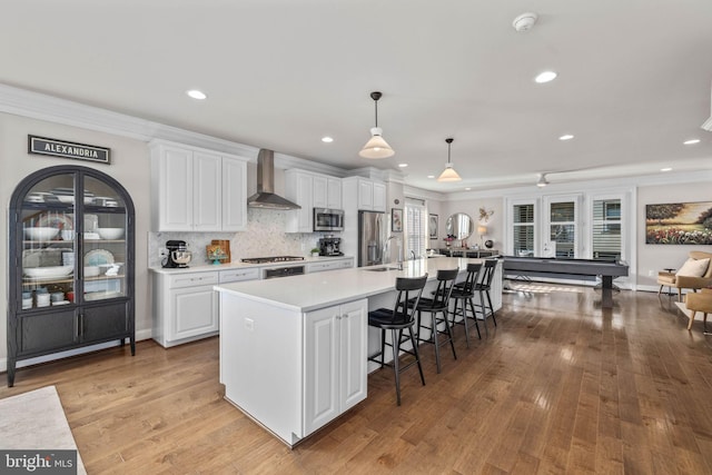 kitchen featuring white cabinetry, hanging light fixtures, a large island with sink, stainless steel appliances, and wall chimney range hood