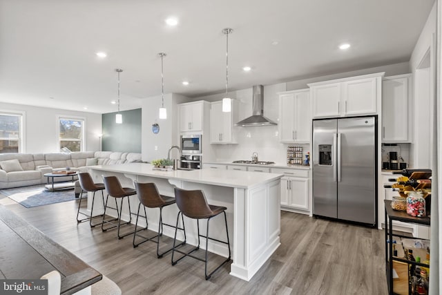 kitchen with wall chimney range hood, stainless steel appliances, an island with sink, and white cabinets