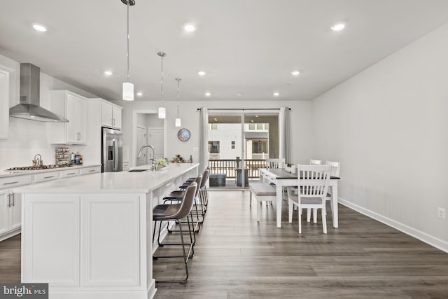 kitchen with sink, wall chimney range hood, appliances with stainless steel finishes, an island with sink, and decorative light fixtures