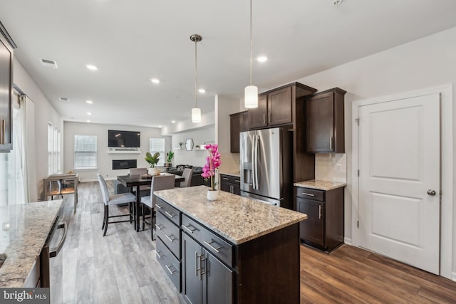 kitchen with dark brown cabinetry, light stone counters, a center island, hanging light fixtures, and stainless steel fridge