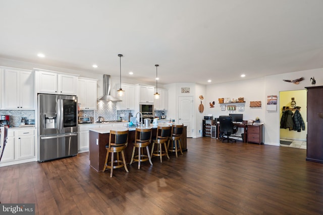 kitchen featuring decorative light fixtures, an island with sink, white cabinets, stainless steel fridge, and wall chimney exhaust hood