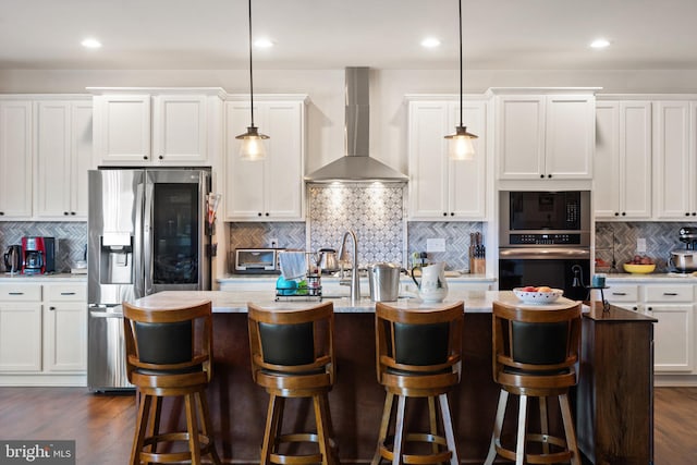 kitchen with stainless steel fridge, wall chimney exhaust hood, decorative light fixtures, and an island with sink