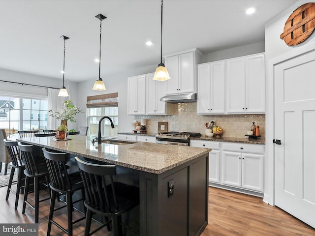 kitchen featuring sink, stainless steel range with gas stovetop, an island with sink, white cabinets, and decorative light fixtures