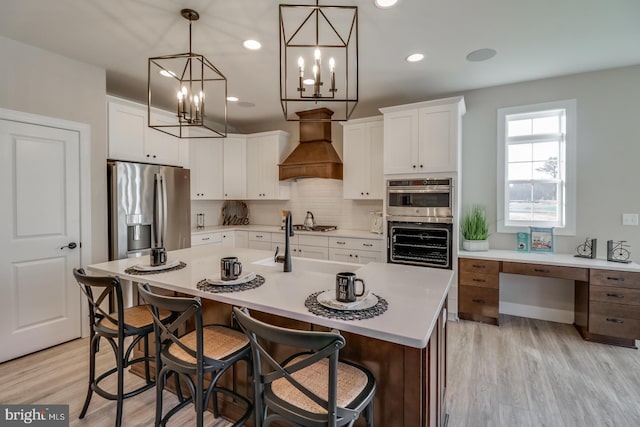 kitchen featuring stainless steel appliances, premium range hood, white cabinets, and an island with sink