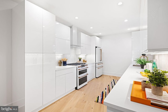 kitchen featuring high end appliances, light wood-type flooring, wall chimney range hood, decorative backsplash, and white cabinets