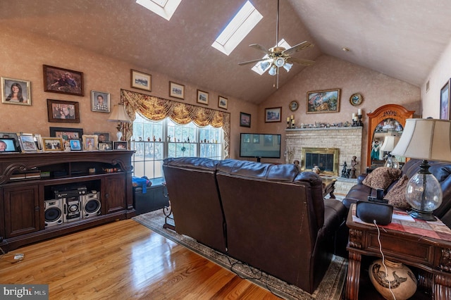 living room with high vaulted ceiling, a skylight, ceiling fan, a brick fireplace, and light hardwood / wood-style flooring