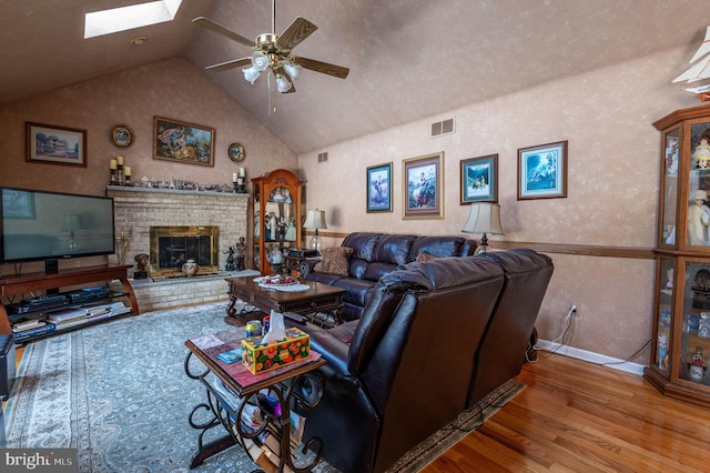 living room featuring ceiling fan, a skylight, high vaulted ceiling, wood-type flooring, and a brick fireplace