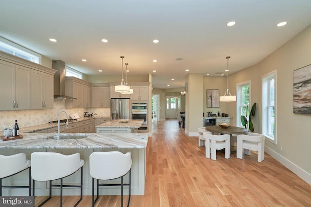 kitchen with appliances with stainless steel finishes, gray cabinetry, hanging light fixtures, light stone counters, and wall chimney exhaust hood