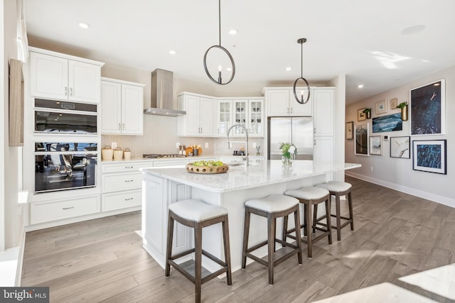 kitchen featuring appliances with stainless steel finishes, white cabinetry, light stone counters, a center island with sink, and wall chimney exhaust hood