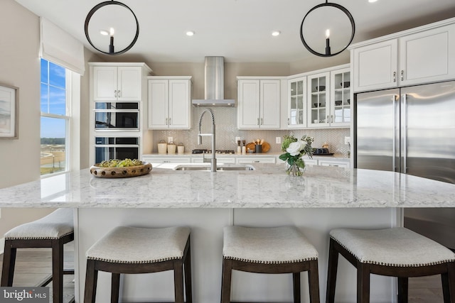 kitchen with wall chimney exhaust hood, high end fridge, white cabinetry, tasteful backsplash, and an island with sink