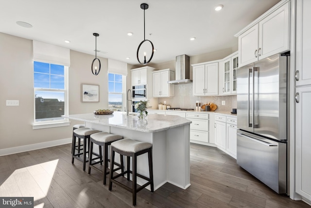 kitchen featuring wall chimney exhaust hood, white cabinetry, appliances with stainless steel finishes, and a center island with sink