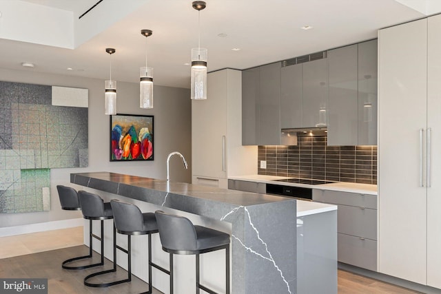 kitchen featuring gray cabinetry, pendant lighting, black electric stovetop, a kitchen island with sink, and decorative backsplash