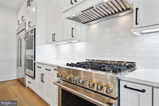 kitchen featuring light stone countertops, custom exhaust hood, stainless steel appliances, white cabinetry, and backsplash