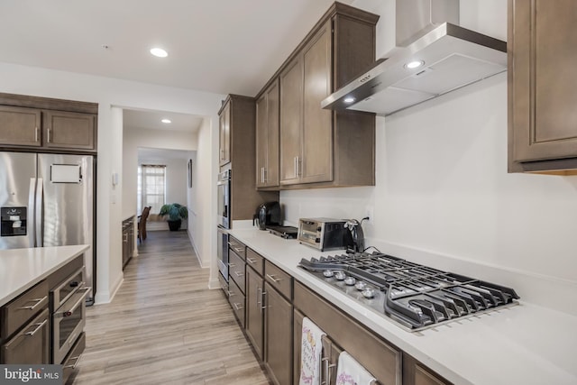 kitchen featuring wall chimney range hood, dark brown cabinetry, light countertops, light wood-style flooring, and appliances with stainless steel finishes