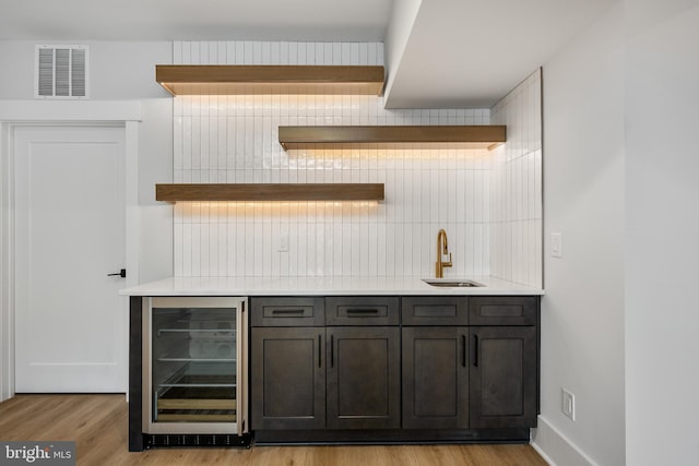 bar featuring sink, wine cooler, backsplash, dark brown cabinets, and light hardwood / wood-style flooring