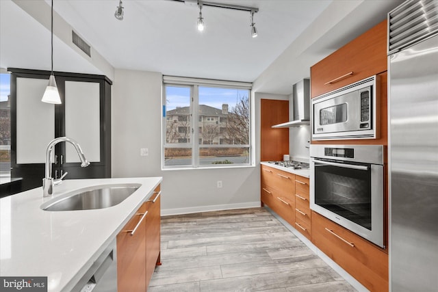 kitchen featuring wall chimney range hood, sink, light hardwood / wood-style flooring, hanging light fixtures, and built in appliances