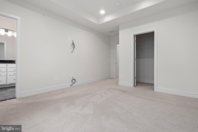 unfurnished bedroom with ensuite bathroom, a spacious closet, a tray ceiling, a closet, and light colored carpet