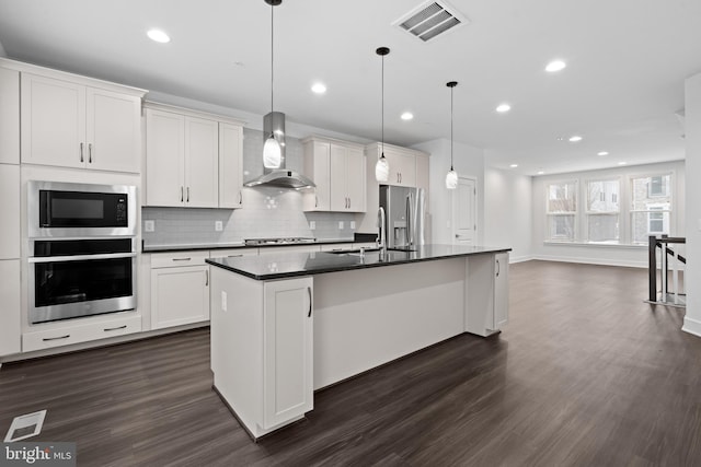 kitchen featuring hanging light fixtures, appliances with stainless steel finishes, an island with sink, wall chimney range hood, and white cabinets