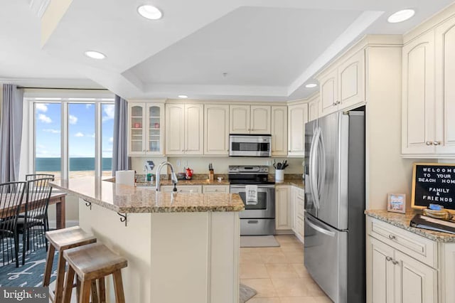 kitchen featuring a breakfast bar, an island with sink, stainless steel appliances, light stone countertops, and a water view