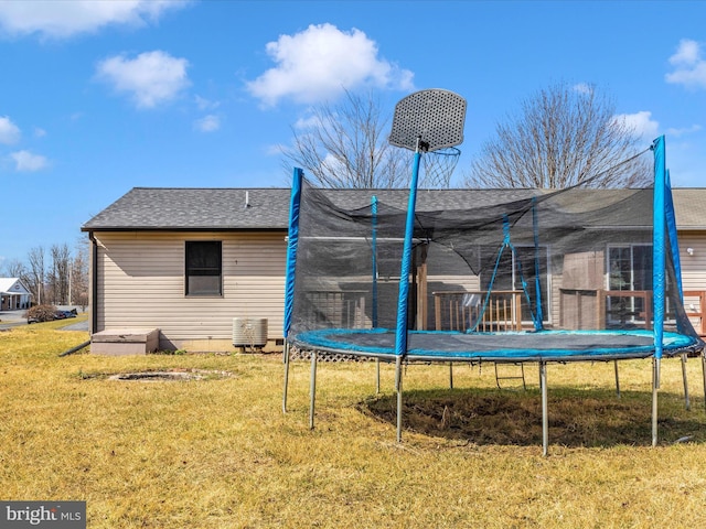 rear view of house with a shingled roof, a trampoline, central AC, and a yard