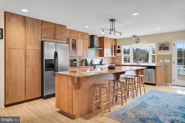 kitchen with a center island, light wood-type flooring, pendant lighting, stainless steel appliances, and wall chimney range hood