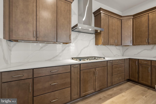 kitchen featuring wall chimney range hood, light hardwood / wood-style flooring, backsplash, light stone countertops, and stainless steel gas stovetop