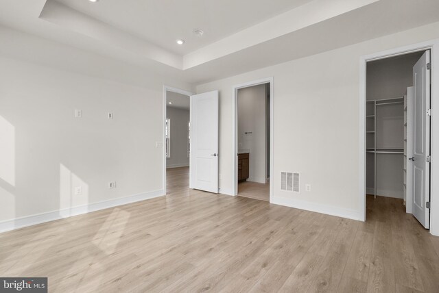 unfurnished bedroom featuring a walk in closet, ensuite bath, a raised ceiling, a closet, and light hardwood / wood-style floors