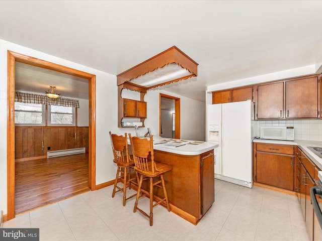 kitchen featuring a breakfast bar, baseboard heating, kitchen peninsula, white appliances, and backsplash