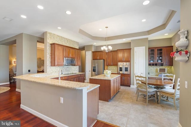 kitchen with pendant lighting, stainless steel appliances, light stone counters, a raised ceiling, and kitchen peninsula