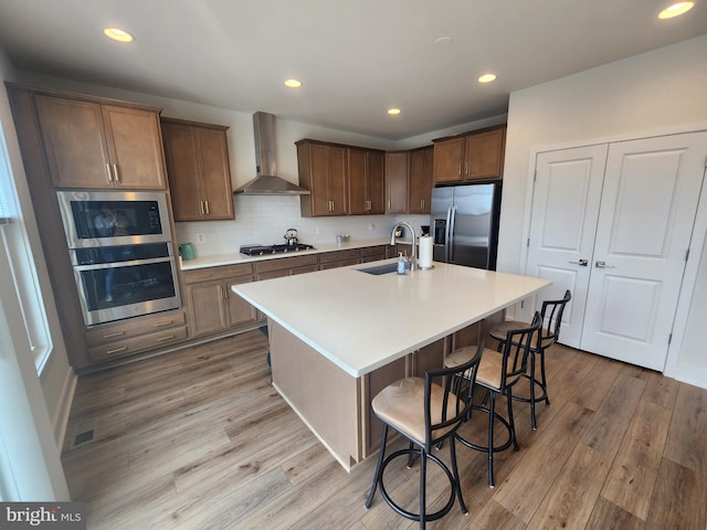 kitchen with stainless steel appliances, light countertops, a sink, an island with sink, and wall chimney exhaust hood