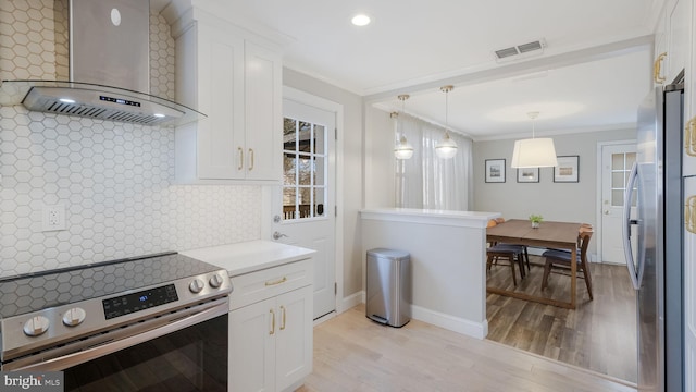 kitchen with ornamental molding, appliances with stainless steel finishes, wall chimney range hood, and white cabinets