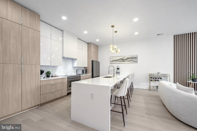 kitchen featuring stainless steel stove, sink, hanging light fixtures, a kitchen island with sink, and light wood-type flooring