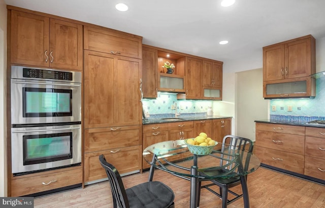 kitchen with sink, dark stone countertops, double oven, tasteful backsplash, and light wood-type flooring