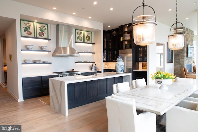 kitchen with wall chimney range hood, a kitchen bar, decorative light fixtures, a large island with sink, and light wood-type flooring