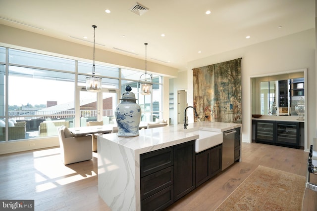 kitchen with sink, hanging light fixtures, a kitchen island with sink, light stone countertops, and light wood-type flooring