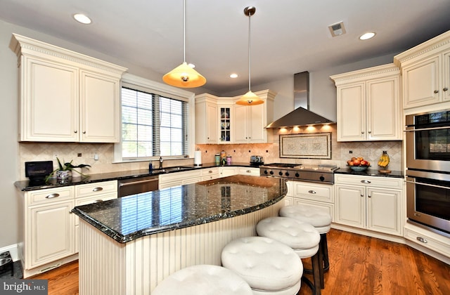 kitchen featuring wall chimney exhaust hood, a kitchen bar, decorative light fixtures, appliances with stainless steel finishes, and a kitchen island