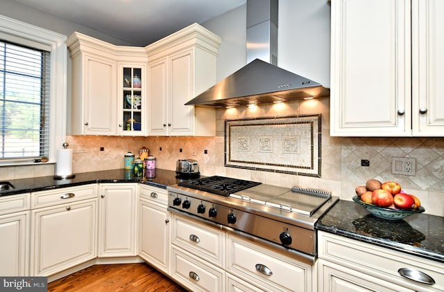 kitchen featuring backsplash, dark stone counters, stainless steel gas cooktop, light hardwood / wood-style floors, and wall chimney exhaust hood