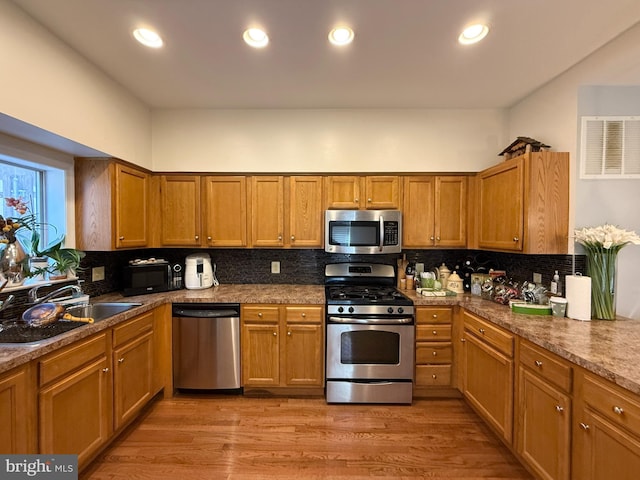 kitchen with brown cabinetry, a sink, stainless steel appliances, light wood-type flooring, and backsplash