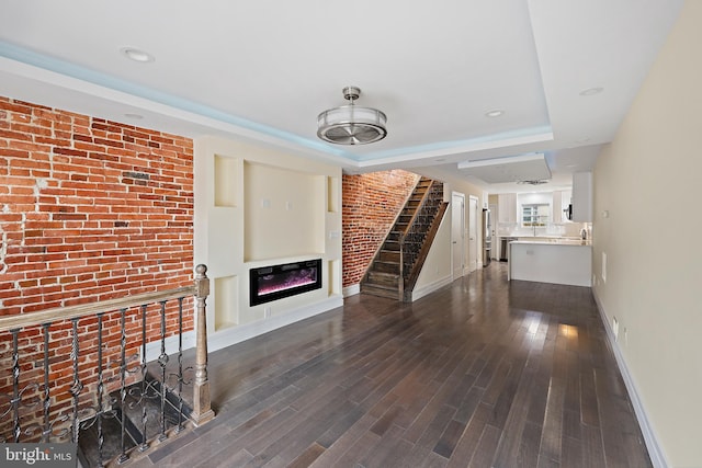 unfurnished living room with dark wood-type flooring, brick wall, and a tray ceiling