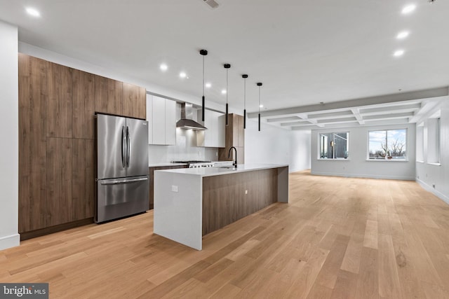 kitchen with wall chimney range hood, appliances with stainless steel finishes, light hardwood / wood-style floors, a center island with sink, and decorative light fixtures