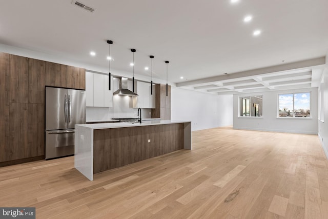 kitchen featuring white cabinetry, hanging light fixtures, a center island with sink, stainless steel fridge, and wall chimney range hood